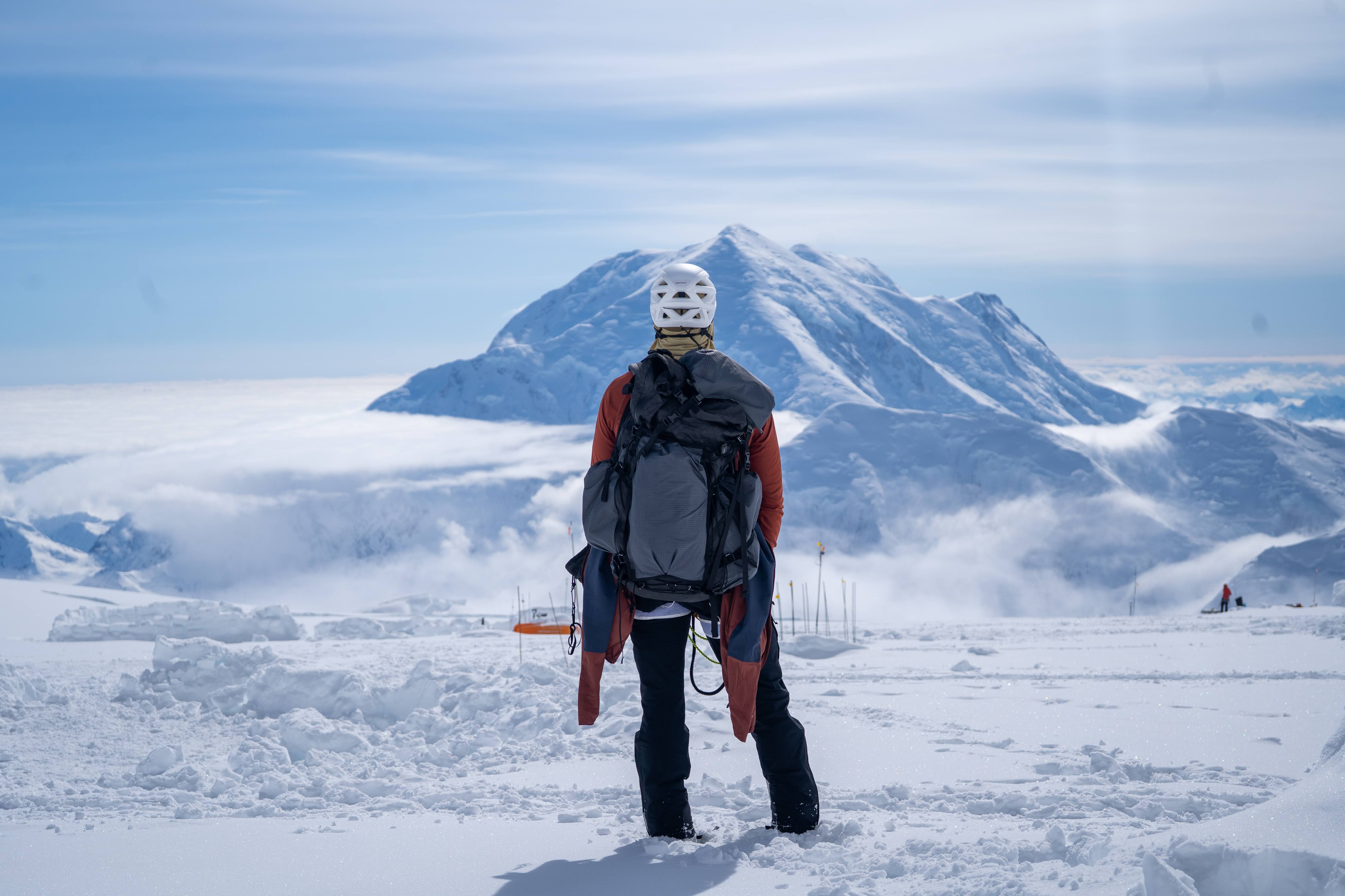 A climber in full gear stands at the top of a snowy peak, with dramatic views of the Denali mountain